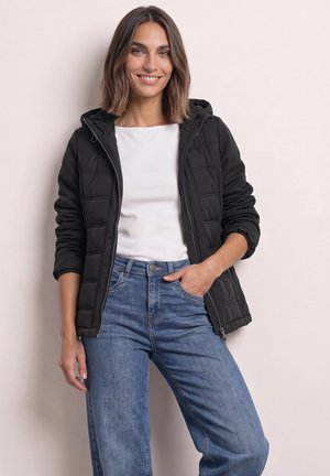 Femme aux cheveux bruns mi-longs portant une veste matelassée noire, une chemise blanche et un jean bleu, souriant avec une main dans la poche.