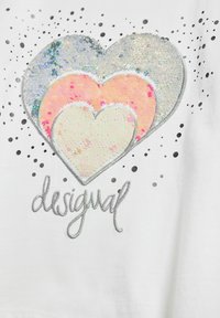 Weißes T-Shirt mit drei geschichteten Herzen aus Pailletten (silber, pink und irisierend), mit verstreuten schwarzen Punkten und "desigual" in Silber gestickt.