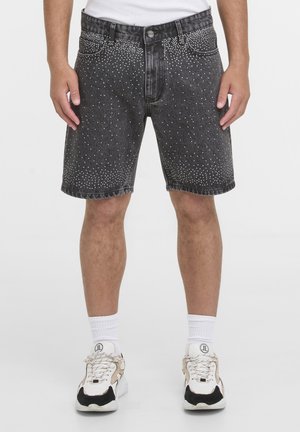 Man die zwarte denim shorts met zilveren studs draagt, witte sokken en witte sneakers met zwarte en beige details.