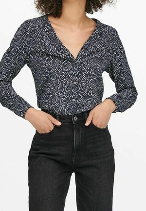 Mujer vistiendo una blusa de manga larga con patrón en blanco y negro, metida dentro de unos vaqueros negros de cintura alta, con las manos en los bolsillos.