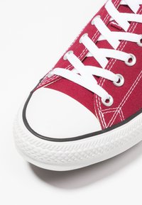 Czerwone płócienne trampki z białym gumowym przodem, czarnym wykończeniem i białymi sznurowadłami. Posiadają metalowe oczka i teksturowaną gumową podeszwę.