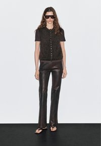 Frau mit dunkler Sonnenbrille, kurzärmeligem Oberteil, flauschiger Knopf-Weste, glänzenden schwarzen Lederhosen und schwarzen Sandalen mit offenen Zehen steht auf schwarzem Boden.