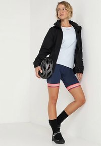 Juoda neperšlampama striukė, šviesiai mėlyna marškinėliai, tamsiai mėlynos dviračių šortai su raudonomis juostelėmis, juodos kojinės ir juoda dviratininko šalmas. Stovinti poza.