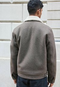Braune Wolljacke mit Schaffellkragen, gerade geschnitten und mit Druckknopfverschluss am Saum. Strukturierter Stoff.