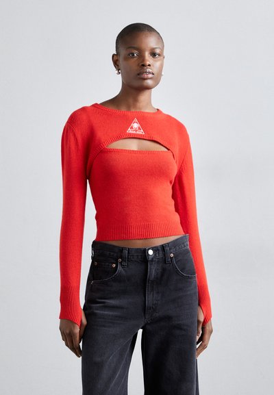 Suéter corto de punto rojo con mangas largas, que presenta una sección recortada y un parche de logo triangular. Combinado con jeans oscuros de tiro alto.