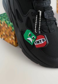 Baskets noires avec un dessus texturé, ornées de tag en caoutchouc vert et rouge représentant des personnages de Minecraft. L'arrière-plan présente des motifs de blocs pixelisés.
