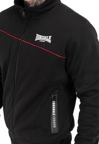 Zwarte zip-up jas met hoge kraag, voorzien van rode accenten langs de borst en een merk-ritszak. Gemaakt van glad, zacht materiaal.