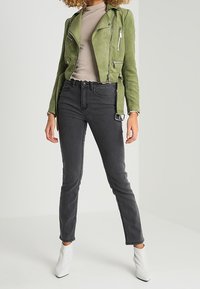 Chaqueta de biker corta de ante verde sobre una blusa beige, combinada con jeans skinny negros y botines blancos, sobre un fondo gris claro.