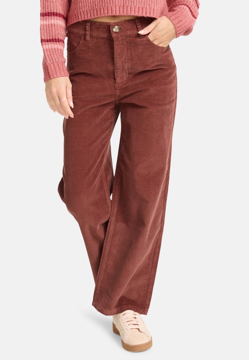 Femme portant un pantalon large en velours côtelé marron taille haute, un pull rayé rose et des baskets rose clair, avec une main dans la poche.