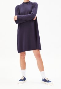 Personne portant une robe longue à manches longues de couleur violet foncé, avec des chaussettes blanches et des baskets bleues, debout les bras croisés sur un fond blanc.