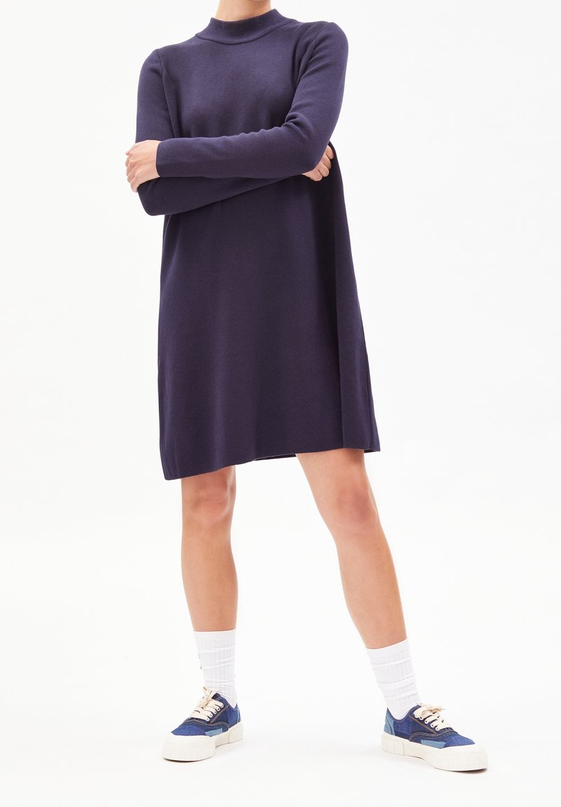 Personne portant une robe longue à manches longues de couleur violet foncé, avec des chaussettes blanches et des baskets bleues, debout les bras croisés sur un fond blanc.