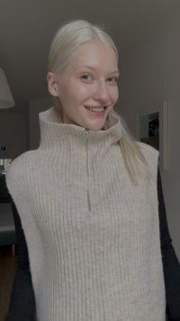 Veste sans manches en maille côtelée beige avec un col haut et une demi-fermeture éclair, portée sur un haut à manches longues noir. Tissu doux et texturé.