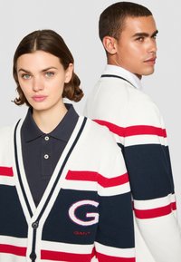 Strikket cardigan i hvitt med marineblå og røde striper, har V-hals, knapplukking og brodert logo på brystet.