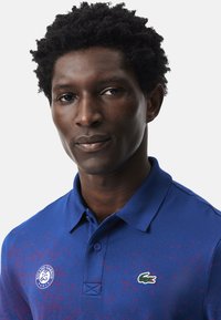 Blå pikétröja med ett strukturerat mönster, prydd med en grön krokodil-logotyp och ett Roland Garros-emblem. Klassisk krage med knäppning.