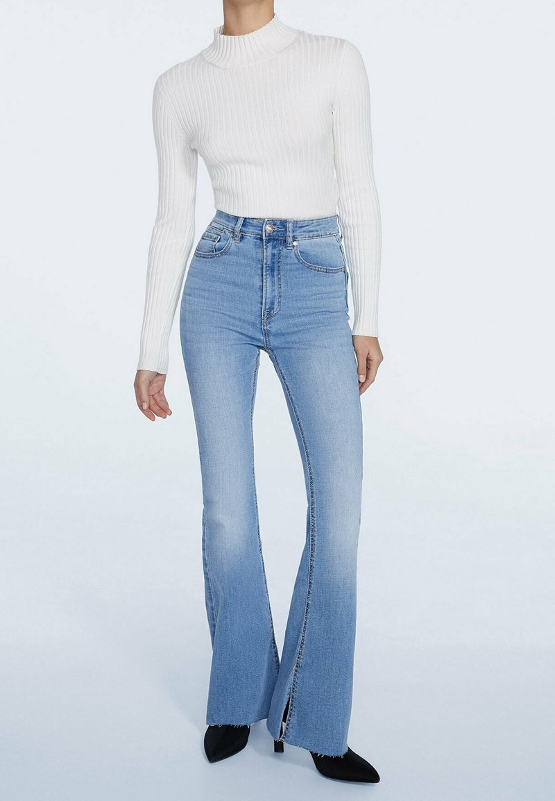 Witte ribgebreide coltrui gecombineerd met lichtblauwe high-waisted flare jeans met voorin uitsparingen en zwarte puntige hakken.