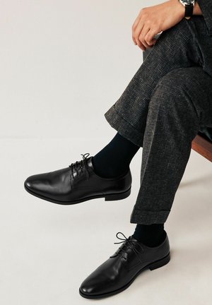 Zapatos de vestir de cuero negro con diseño de cordones, puntera redonda y un acabado suave. Llevados con calcetines oscuros y pantalones grises con estampado.