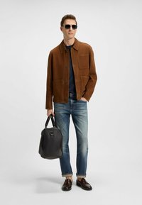 Giacca in suede marrone con due tasche frontali, camicia blu navy sotto, jeans blu chiaro con orlo risvoltato, borsa in pelle nera e occhiali da sole tondi.