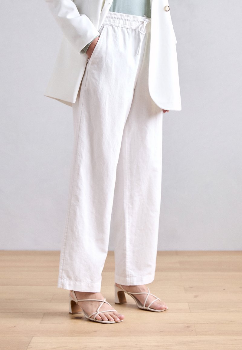 Personne portant un pantalon blanc à jambes larges avec une taille élastique, un blazer blanc et des sandales beiges à talons et à brides, se tenant debout sur un sol en bois.