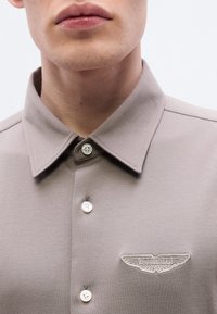 Πόλο μπλουζάκι σε χακί χρώμα με δομημένο γιακά, δύο λευκά κουμπιά και κεντημένο λογότυπο Aston Martin στο στήθος.