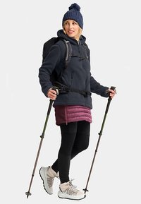 Fleecejack in marineblauw, gedragen over een bordeauxkleurige geïsoleerde rok, zwarte legging en lichte wandelschoenen. Met wandelstokken en een rugzak.