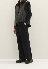 Schwarze Lederweste mit einem Fellkragen, Reißverschluss vorne und Seitentaschen, kombiniert mit schwarzen weiten Cargo-Hosen und schwarzen Schuhen.