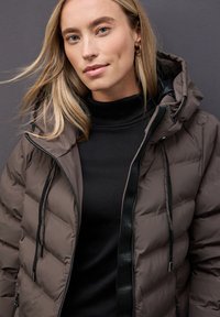 Hnědá péřová bunda s kapucí, která má prošívanou texturu, černý zip a stahovací šňůrky. Nositá přes černý rolák.