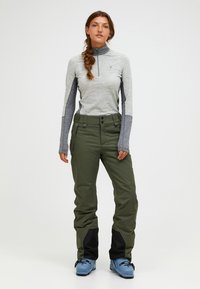 Grijze lange mouwen zip-up top gecombineerd met olijfgroene skibroeken met zwarte onderbenen en blauwe skischoenen. Gladde stof, slim fit.