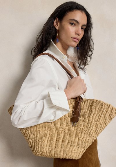 Sötét göndör hajú nő fehér blúzt és színes fülbevalókat visel, kezében nagy, barna bőrpántos, fonott szalma táska.