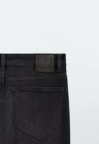Jean en denim noir avec une texture lisse, dotés d'une étiquette en cuir à la taille et d'une poche arrière au design simple.