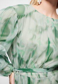 Průhledné zelené šaty s měkkými sklady, oversized, nařasenými rukávy a látkovým páskem. Materiál vykazuje vzor ve stylu akvarelu.