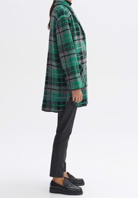 Frau trägt einen grün-schwarz karierten Mantel, schwarze Hosen und schwarze Slipper, steht im Profil vor einem weißen Hintergrund.