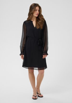 Femme portant une robe noire mi-longue avec des manches longues transparentes, une ceinture à la taille et des sandales noires, debout devant un fond clair uni.