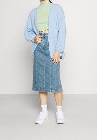 Osoba oblečená v světle modrém kardiganu, bledě zeleném zkráceném roláku, džínové midi sukni, bílých ponožkách a bílých teniskách, stojící na jednobarevném pozadí.