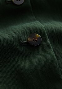 Tissu vert foncé avec un gros plan sur un bouton sombre et brillant à quatre trous cousu, mettant en valeur la texture du tissu et les détails de la couture.