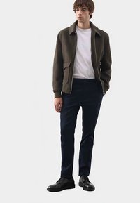 Men's olive green bomber jacket with a white T-shirt, dark blue pants, and black shoes. Features front pockets and a zip closure.