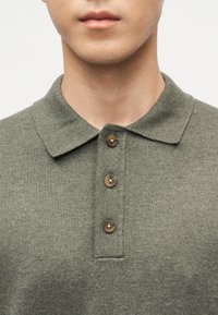 Άνδρας που φοράει πράσινο πλεκτό πόλο πουκάμισο με γιακά και τρία καφέ κουμπιά στην πατιλέτα, εμφανιζόμενος από τον λαιμό έως το στήθος.