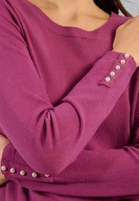 Maglione viola a maniche lunghe con bottoni perle sui polsini indossato da una persona con le braccia incrociate.