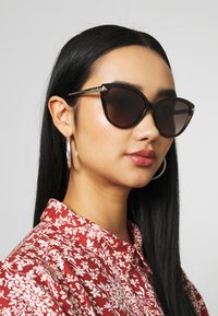Ochelari de soare cu formă cat-eye, cu lentile închise, rame cu model țestoasă și detalii aurii. Purtați cu o cămașă roșie cu model floral.