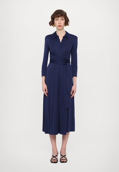 Mornarsko plava haljina na preklop s dugim rukavima, kopčanjem na gumbe sprijeda i pojasom u struku, s glatkom teksturom i lepršavom suknjom do gležnjeva. Crne sandale s remenčićima.