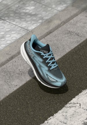 Light blue and black Hoka running shoe resting on textured pavement near a tactile pedestrian curb ramp and crosswalk markings.
