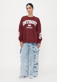 Sweatshirt carmesim com impressão de "DETROIT" e "MICHIGAN", combinado com umas calças de carga oversized de cor azul-claro com múlticos bolsos e fechos éclair.