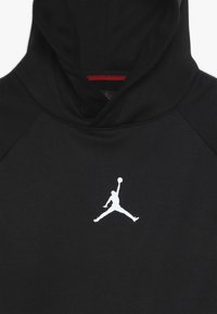 Černá mikina s kapucí vyrobená z hladkého materiálu, s bílým logem Jumpman na hrudi a červeným akcentem na vnitřní straně kapuce.