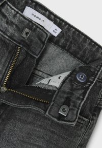 Jeans en denim gris foncé avec une fermeture à glissière et un bouton, présentant une texture lisse et des coutures contrastantes au niveau de la taille.