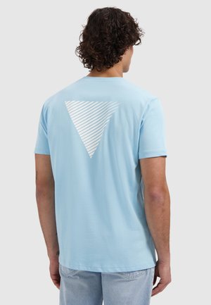 Mann mit lockigem Haar, der ein hellblaues T-Shirt mit einem weißen, diagonal gestreiften Dreieck auf dem Rücken und hellblaue Jeans trägt, von hinten gesehen.