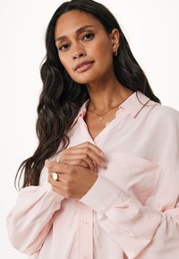 Blusa lunga manica color rosa chiaro con bottoni frontali, taschino sul petto e texture morbida. Modella che indossa orecchini a cerchio dorati e un anello, mani incrociate.