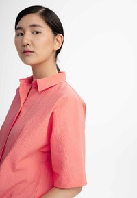 Korte mouwen shirt van textuurstof in koraalkleur, met een klassieke kraag en knoopdetails, gepresenteerd tegen een effen witte achtergrond.