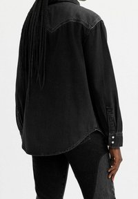 Une personne avec de longs cheveux tressés porte une chemise en denim noir ample et un pantalon noir, vue de dos devant un fond uni.