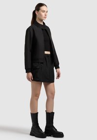 Femme aux longs cheveux attachés en queue de cheval, portant un haut noir court, une veste bomber noire, une mini-jupe noire avec poche et des bottes de combat noires à lacets.