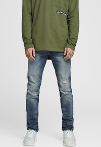 Man som bär urblekta blå slitna jeans, vita sneakers och en olivgrön långärmad skjorta med bröstficka och logopatch.