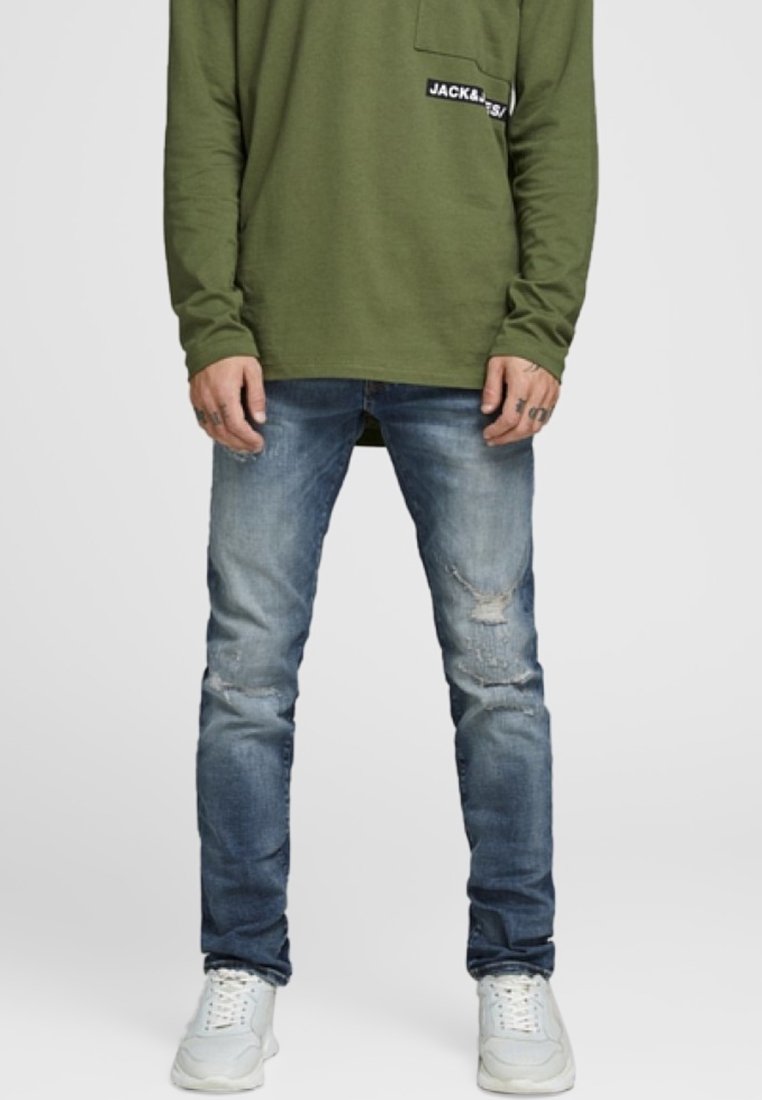 Man som bär urblekta blå slitna jeans, vita sneakers och en olivgrön långärmad skjorta med bröstficka och logopatch.